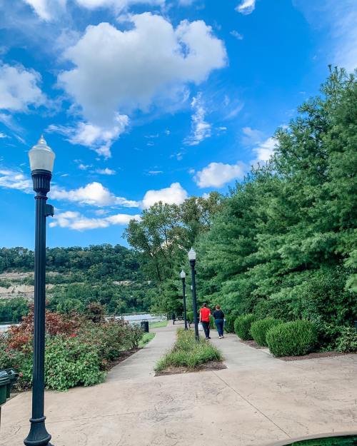 A scenic pathway lined with trees and lampposts, with two people walking under a blue sky with fluffy clouds.