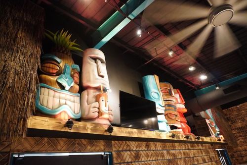 Colorful tiki statues displayed on a wooden shelf in a tropical-themed interior with a ceiling fan above.