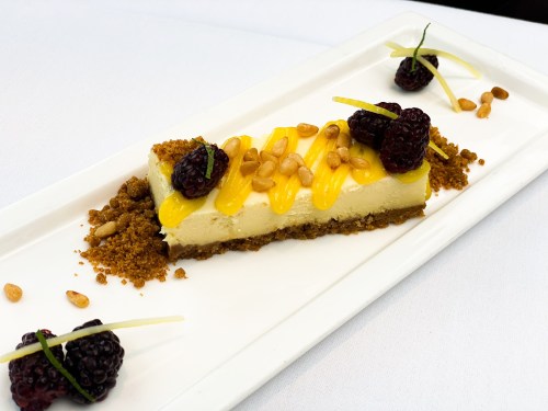 A slice of cheesecake topped with mango puree, blackberries, pine nuts, and garnished with lime zest on a white plate.