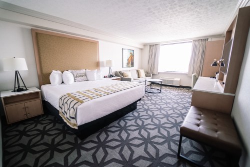 A modern hotel room featuring a king-sized bed, seating area, desk, and large window with natural light.