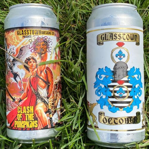 Two beer cans on grass: one features a warrior with mythical creatures, the other has a decorative crest and "October" label.