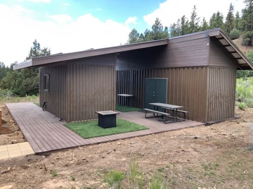 A modern brown cabin with a wooden deck, surrounded by trees and open space, featuring outdoor tables.