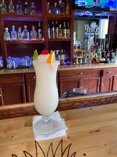 A tropical cocktail with fruit garnishes sits on a bar counter, surrounded by bottles of liquor in the background.