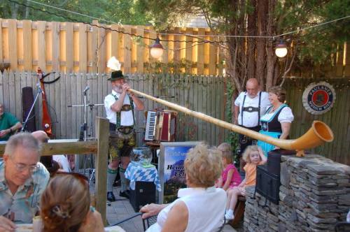 A musician in traditional attire plays a long horn at an outdoor gathering, with guests enjoying the performance.