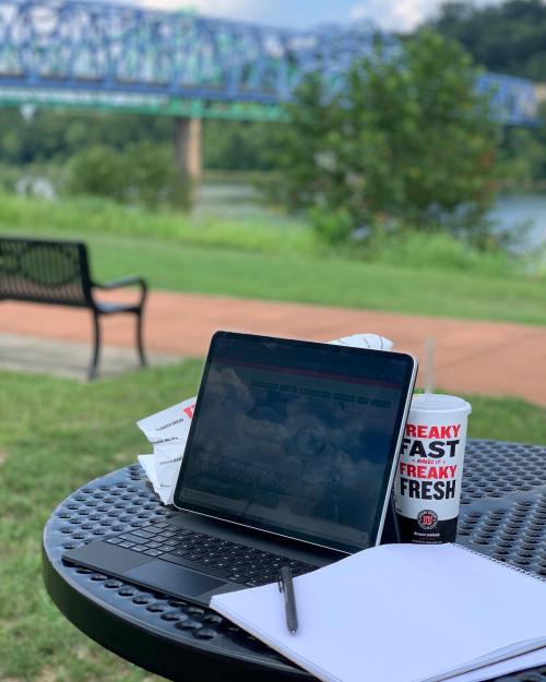 A laptop on a table with a drink, overlooking a river and bridge, with a notebook and pen nearby.