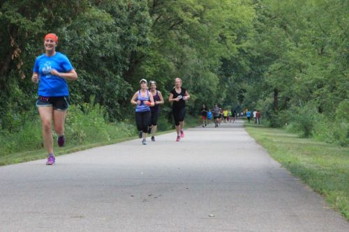 A group of runners on a tree-lined path, some smiling and others focused, enjoying a sunny day outdoors.