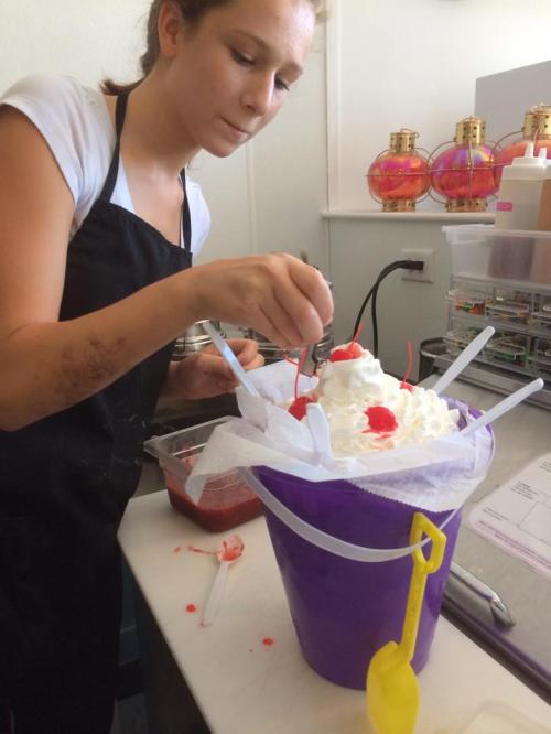A young girl in an apron decorates a large ice cream sundae with whipped cream and cherries in a shop setting.