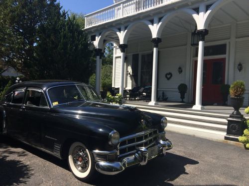 A classic black car parked in front of a charming white house with columns and a red door.
