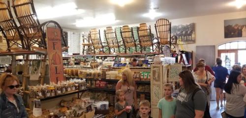 A busy market interior filled with shelves of goods, people browsing, and wooden chairs displayed overhead.
