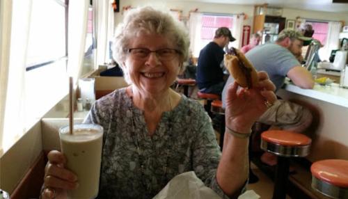A smiling older woman holds a milkshake in one hand and a sandwich in the other, sitting in a diner.