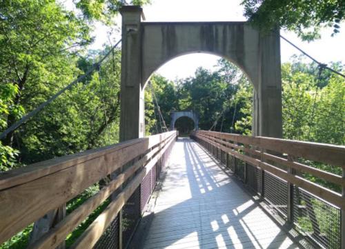 A wooden suspension bridge with a stone arch, surrounded by lush green trees and dappled sunlight.