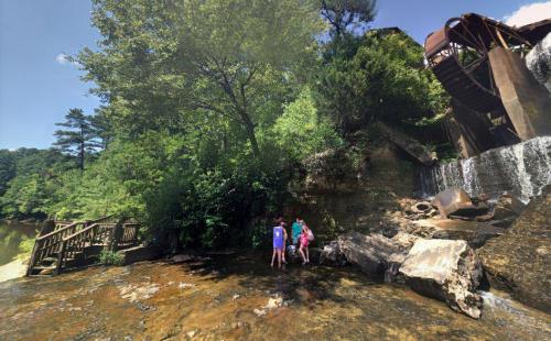 Two children play near a rocky riverbank, with lush greenery and a watermill in the background under a clear blue sky.