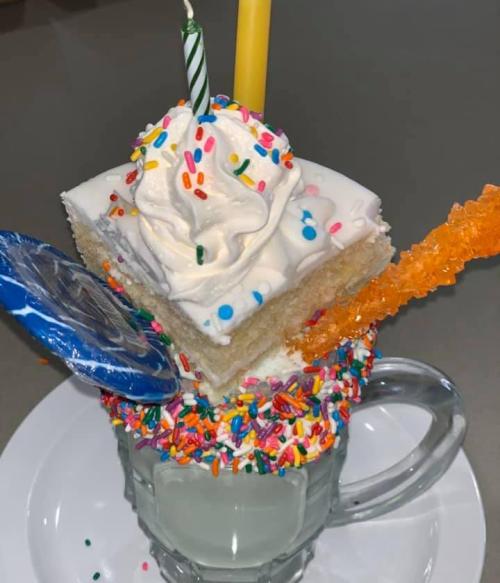 A colorful dessert with a cake slice topped with whipped cream, sprinkles, and a candle, served in a glass mug.