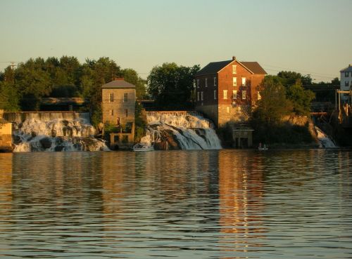 A serene view of a waterfall beside historic buildings, reflecting in calm water under a golden sunset.