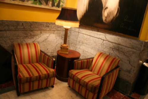 Two striped armchairs beside a round table and a lamp, set against a marble wall and a horse painting.