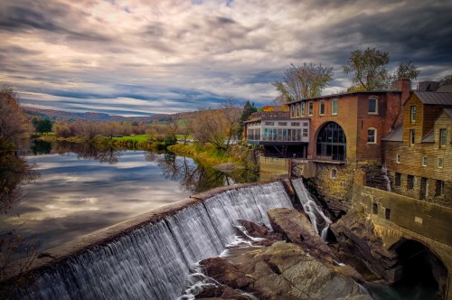 A scenic view of a dam with a waterfall, surrounded by trees and a cloudy sky, reflecting in the calm river below.