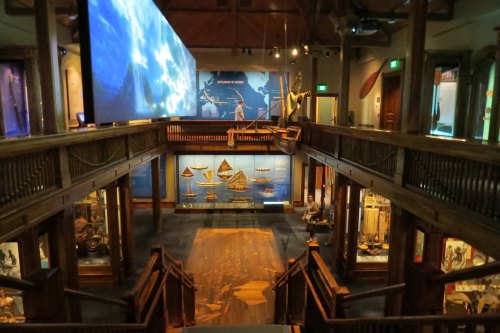 Interior view of a museum showcasing boats and maritime exhibits, with a large screen displaying ocean imagery above.