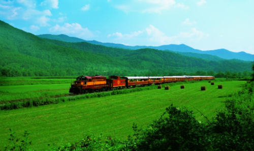 A colorful train travels through a lush green landscape with mountains in the background under a clear blue sky.