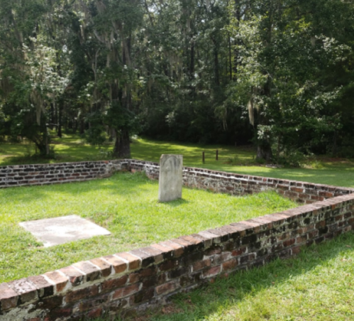 A small, grassy cemetery enclosed by a brick wall, featuring a weathered gravestone under a canopy of trees.