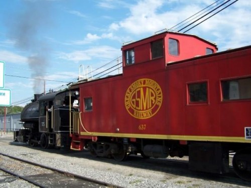 A vintage steam locomotive and red caboose parked on a railway track under a blue sky.