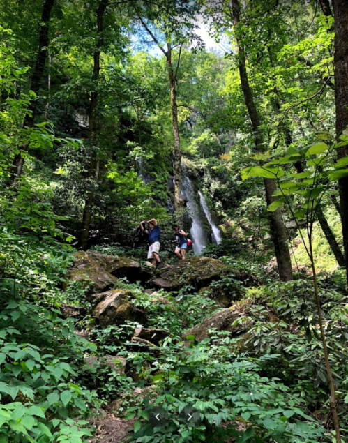 Two hikers ascend a rocky path through lush greenery, with a waterfall visible in the background.