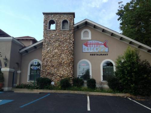 Exterior of a restaurant with a stone tower, large fish logo, and "OPEN" sign, surrounded by greenery and parking spaces.