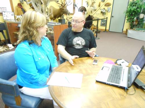 Two people sit at a table in an office, engaged in conversation, with a laptop and notes in front of them.
