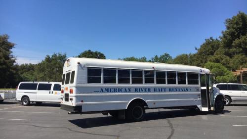 A white bus labeled "American River Raft Rentals" parked in a lot with trees and other vehicles in the background.