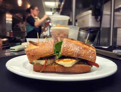 A close-up of a sandwich with grilled vegetables and cheese, served on a plate in a kitchen setting.
