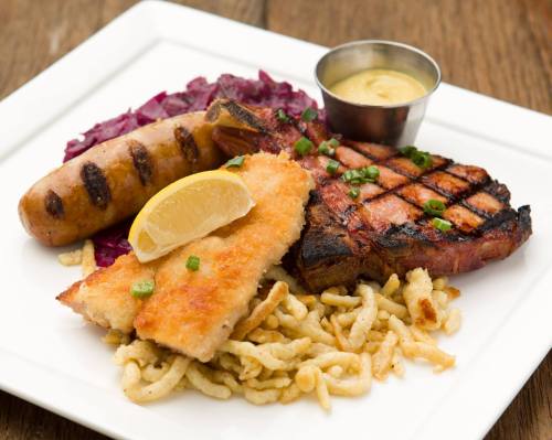 A plate featuring grilled sausage, pork chop, breaded fish, and red cabbage, garnished with a lemon wedge and sauce.