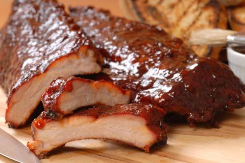 Close-up of tender, glazed barbecue ribs on a wooden cutting board, with a side of sauce and grilled items in the background.