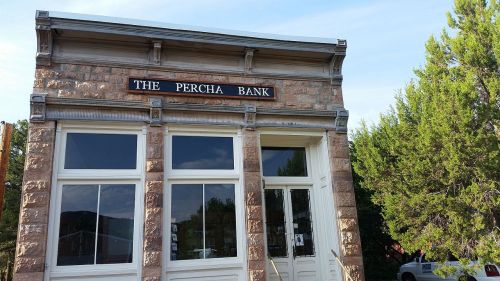 Historic stone building with large windows, featuring a sign that reads "The Percha Bank." Green trees in the background.