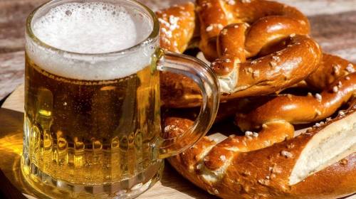 A frosty mug of beer next to a plate of golden-brown pretzels on a wooden surface.