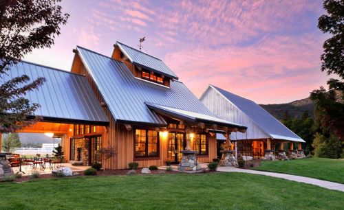 A modern wooden house with a metal roof, surrounded by greenery, under a colorful sunset sky.