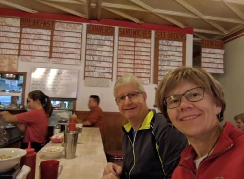 A man and woman smile at the camera while sitting at a diner counter with a menu board in the background.
