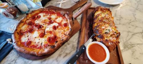 A pepperoni pizza on a wooden board next to a cheesy garlic bread with marinara sauce on a marble table.