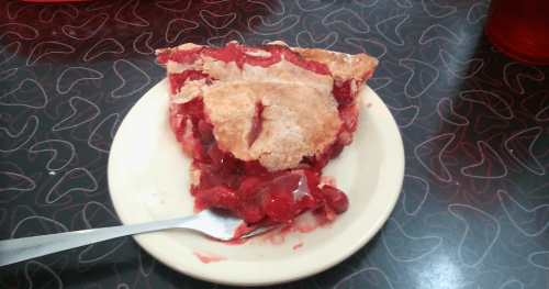 A slice of cherry pie on a plate, with a fork beside it, sitting on a patterned table.