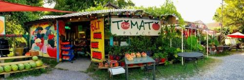 A colorful market stand named "The Tomato Place" surrounded by greenery, featuring fresh produce and vibrant decorations.