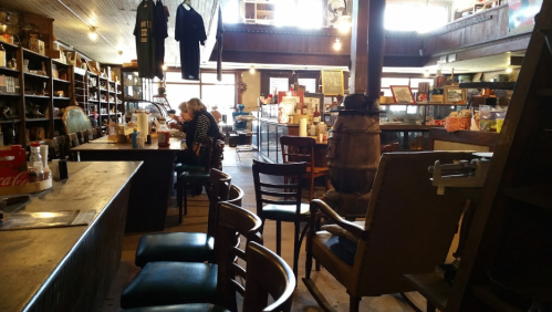 A cozy café interior with wooden furniture, shelves filled with items, and a few patrons enjoying their time.
