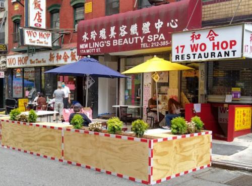 Outdoor dining area with umbrellas at Wo Hop Restaurant, surrounded by planters on a busy street in Chinatown.