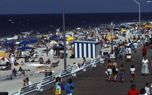 Crowded beach scene with colorful umbrellas, people sunbathing, and a boardwalk along the shore.
