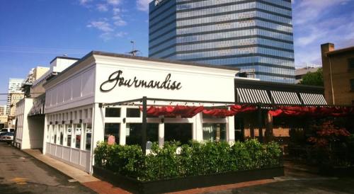 Exterior of a restaurant named "Gourmandise," featuring a white facade and outdoor seating, with a city skyline in the background.