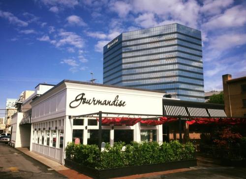 A modern restaurant named "Gourmandise" with outdoor seating, set against a blue sky and tall buildings in the background.