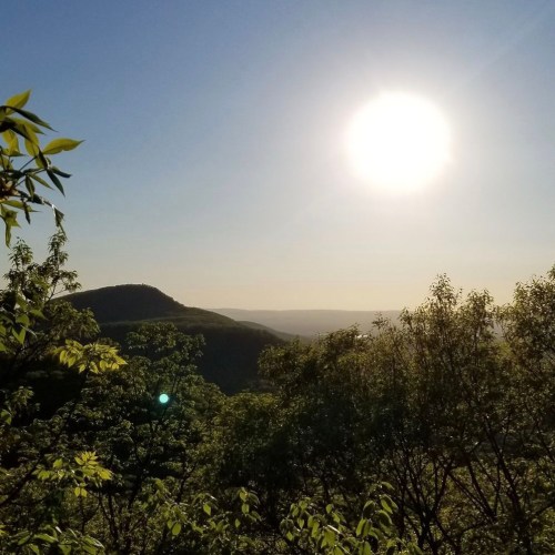 A scenic view of mountains under a bright sun, surrounded by lush greenery and a clear blue sky.