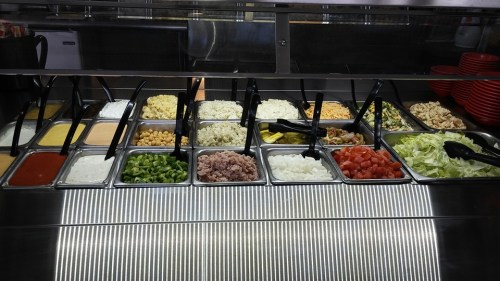 A variety of fresh toppings and sauces displayed in a salad bar, including vegetables, grains, and dressings.