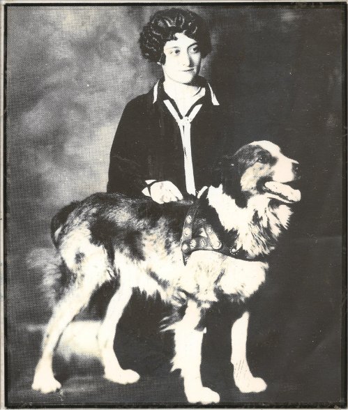 A vintage black-and-white photo of a young woman in a dark outfit standing beside a large dog.