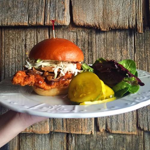 A hand holding a crispy chicken sandwich with coleslaw, barbecue sauce, and pickles, served with a side salad.