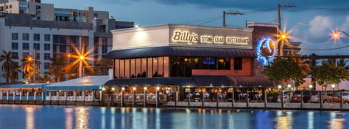 A waterfront restaurant, "Billy's Stone Crab," illuminated at dusk, with outdoor seating and palm trees nearby.