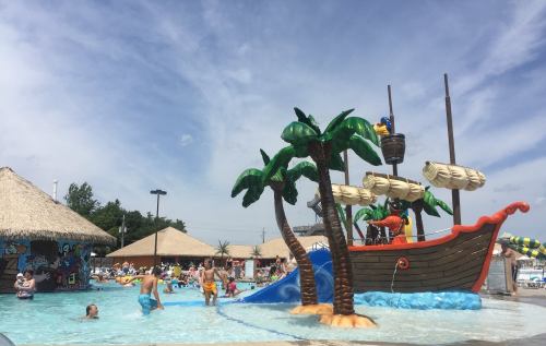 A vibrant water park scene with a pirate ship, palm trees, and children playing in the pool under a blue sky.