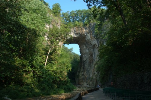 A natural stone arch surrounded by lush green trees and a pathway, creating a serene outdoor scene.